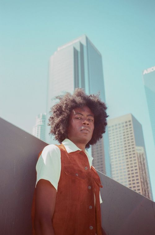 An individual with an afro stands in front of city skyscrapers, wearing a corduroy vest and collared shirt, against a light blue sky.