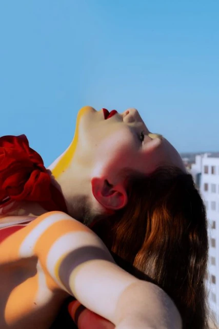 Adult female with red lipstick is tilted back looking at the bright blue sky with buildings blurred in the background in an outdoor setting.
