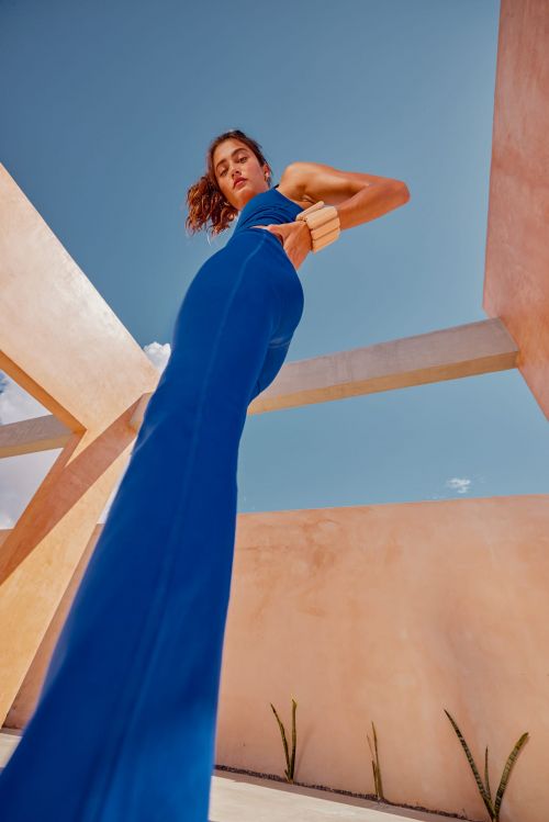 Woman in blue jumpsuit poses against blue sky and architectural structures, captured from a low angle.