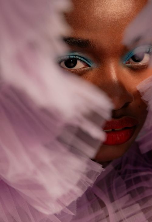 Close-up portrait of a Black woman with bright blue eyeshadow and red lipstick framed by soft lavender tulle fabric.