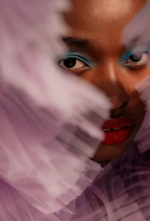 Close-up portrait of a Black woman with bright blue eyeshadow and red lipstick framed by soft lavender tulle fabric.