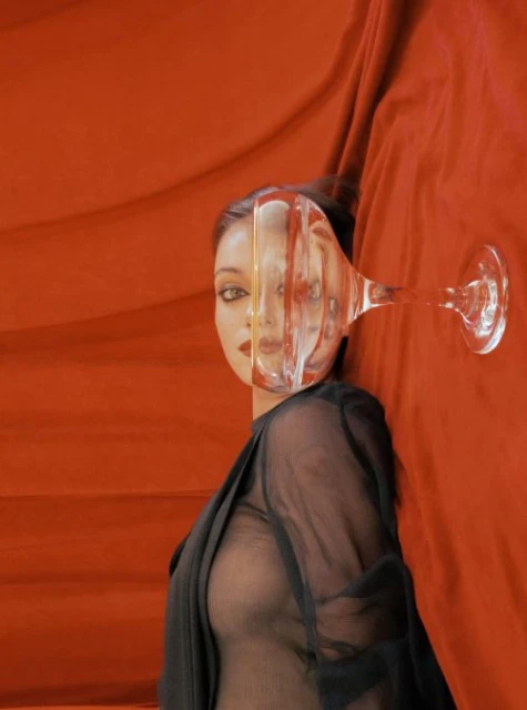 Surreal portrait of a woman's face partially distorted through a transparent glass, set against a bold orange fabric backdrop.
