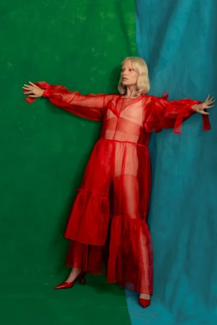 A blonde female fashion model poses in a see-through red dress and red heels against a background divided into green and blue colors.