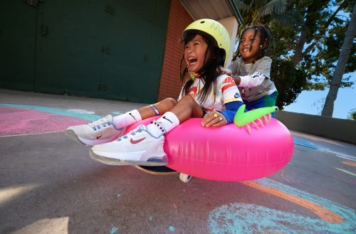 Two children, one wearing a helmet, sit on a pink inflatable tube on a skateboard, on concrete with colorful chalk drawings in an outdoor setting.