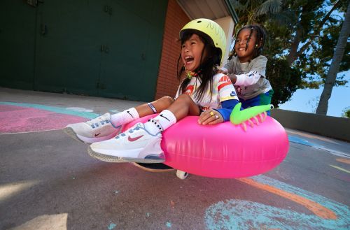 Two young children, one wearing a helmet, smile and laugh while riding on an inflatable pink tube outside with trees and a building behind them.