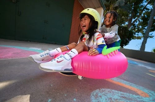 Two young children, one wearing a helmet, smile and laugh while riding on an inflatable pink tube outside with trees and a building behind them.