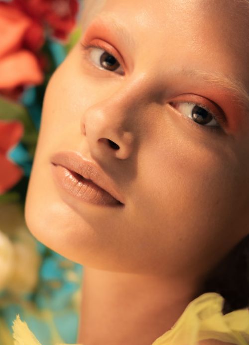 Close-up portrait of a woman with orange eyeshadow, glossy lips, and light hair surrounded by vibrant flowers and soft yellow fabric.