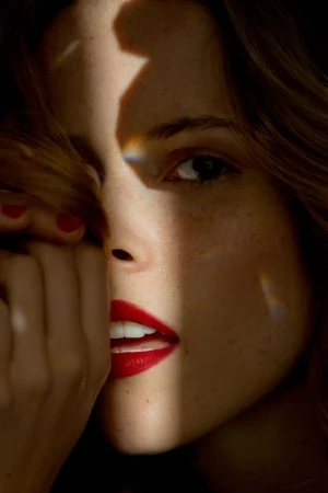 Close up portrait of a woman with red lipstick and freckles, partially obscuring her face with her hand.