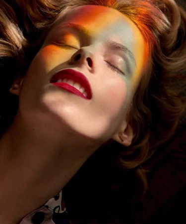 Close-up portrait of a woman with vibrant prismatic light projected across her face and closed eyes, wearing bright red lipstick.