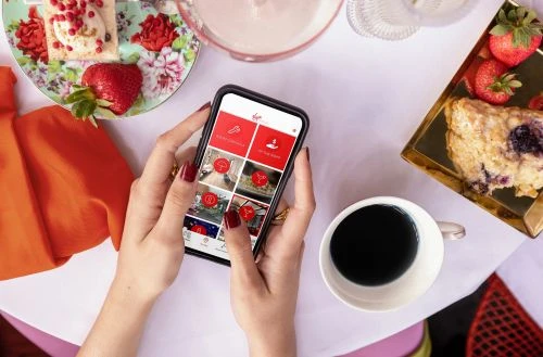 Overhead shot of a person holding a smartphone with a food photography app screen, surrounded by pastries, coffee, and fresh strawberries.