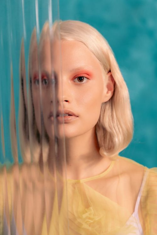Close-up portrait of a blonde woman with bright coral eye makeup, partially obscured by a textured glass pane against a teal backdrop.