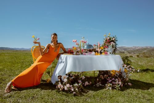 Woman in an orange dress poses with flowers at a decorated table in a grassy field under a blue sky, with a distant mountain view.