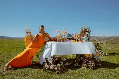 Woman in an orange dress poses with flowers at a decorated table in a grassy field under a blue sky, with a distant mountain view.