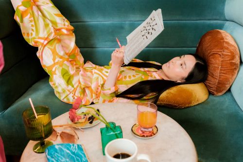 Asian woman in patterned clothing reads newspaper on blue couch. Drinks and snacks on marble table in interior shot. 
