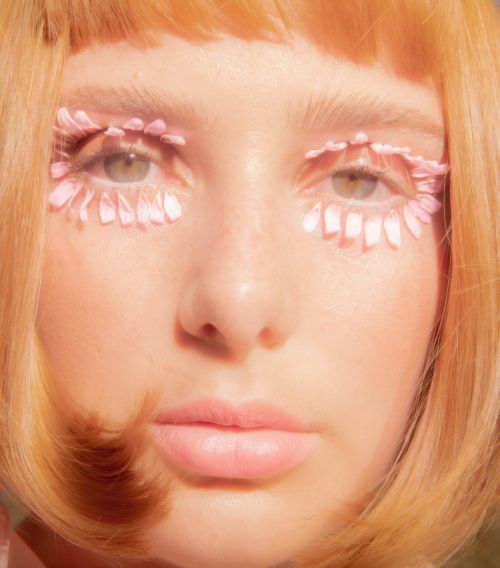 Close-up of a young woman with light skin, red hair, and pink flower petals arranged around her eyes in a creative beauty shot.