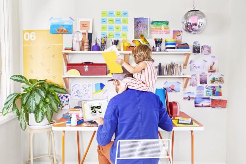 Father works at desk with laptop while daughter sits on his shoulders reading a book in bright, modern, home office interior.