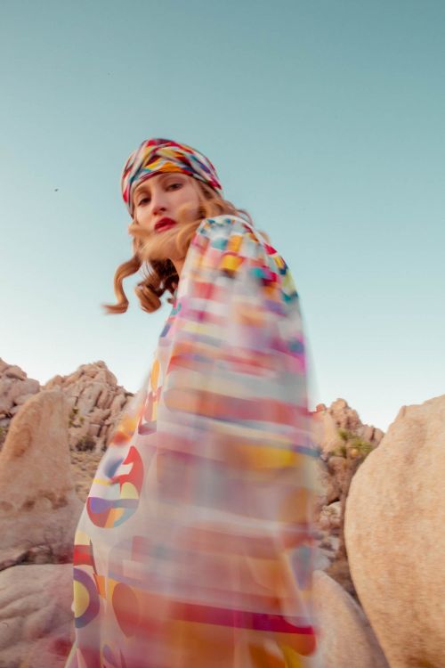 Woman in a patterned headscarf and dress poses among rock formations under a clear sky in an outdoor setting, fashion photography.