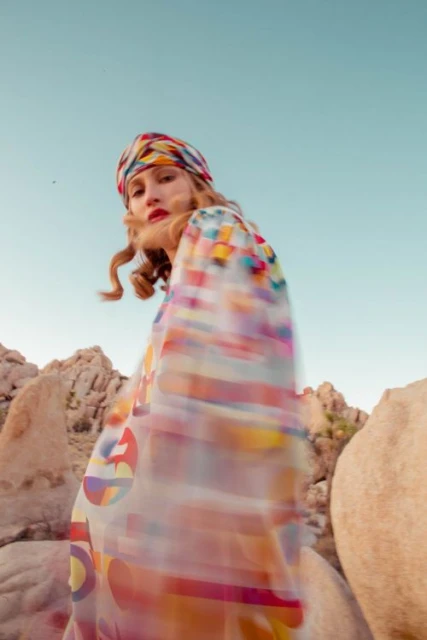 Woman in a patterned headscarf and dress poses among rock formations under a clear sky in an outdoor setting, fashion photography.