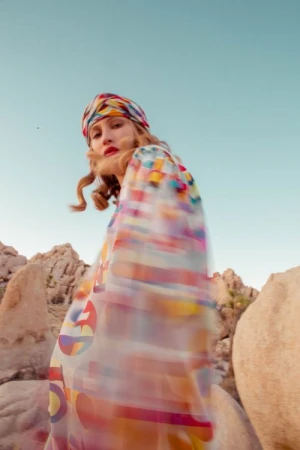 Woman in a patterned headscarf and dress poses among rock formations under a clear sky in an outdoor setting, fashion photography.