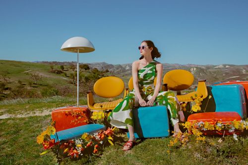 Young woman in a leaf-print jumpsuit sitting on colorful vintage luggage outdoors in a grassy field under a clear blue sky.