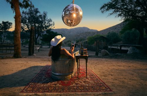 Woman in a cowboy hat relaxes in a metal tub with wine under a disco ball in a desert setting at dusk, creating a vintage aesthetic.