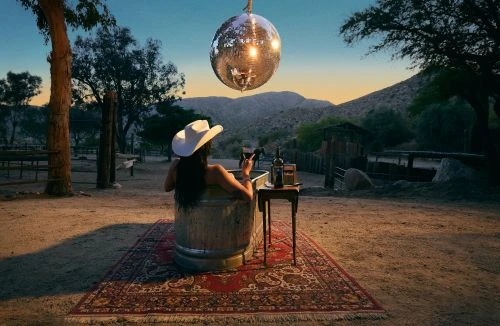Woman in a cowboy hat relaxes in a metal tub with wine under a disco ball in a desert setting at dusk, creating a vintage aesthetic.