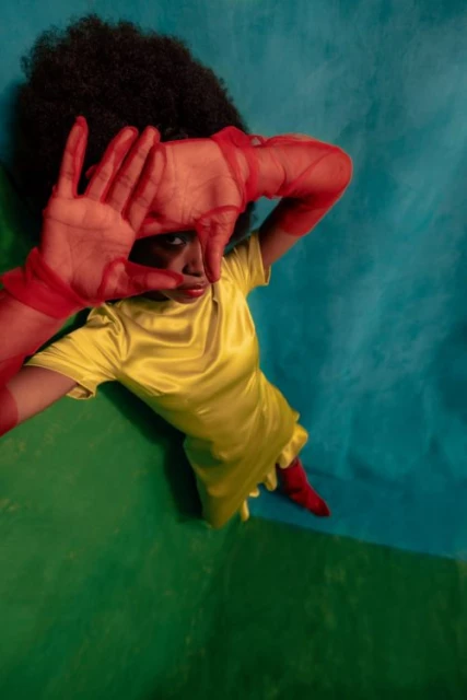 Stylish portrait of a woman with an afro, wearing a yellow dress and red gloves, playfully framing her face against a blue and green backdrop.