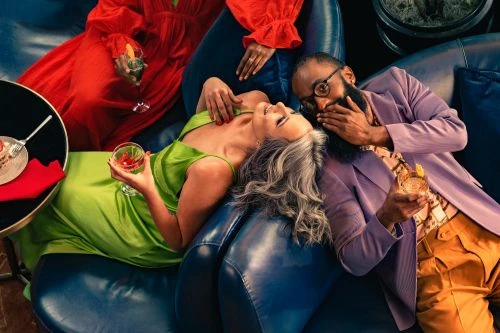 Overhead shot of three people lounging on a blue sofa, two holding cocktails, with one covering his mouth in surprise.