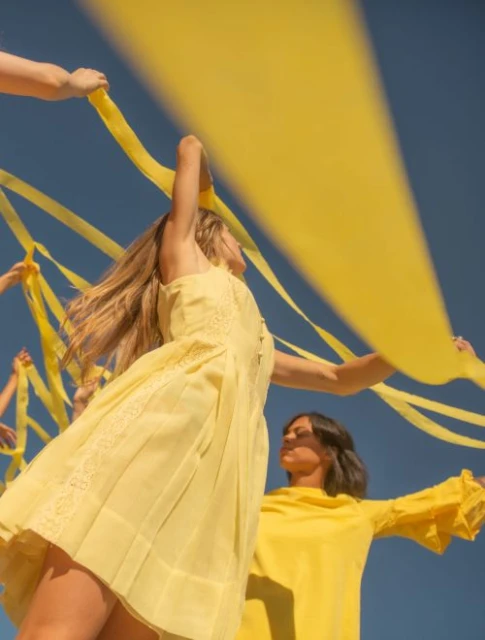 Two women in yellow dresses hold yellow ribbons against a blue sky, illuminated by sunlight, creating a vibrant and sunny visual.
