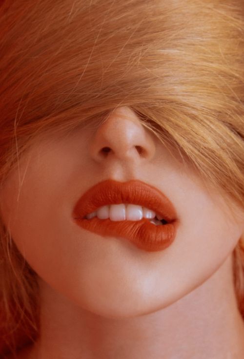 Close-up of a woman's face with red lipstick, fair skin, and blonde hair partially covering her eyes, in a beauty or fashion context.