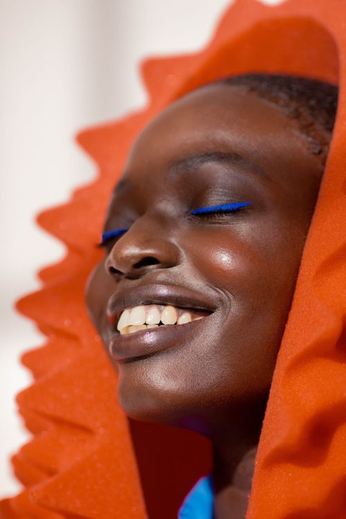 Portrait of a smiling Black woman with vibrant blue eyeliner, wearing a textured orange hood, in soft natural light.
