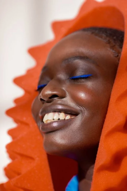 Portrait of a smiling Black woman with vibrant blue eyeliner, wearing a textured orange hood, in soft natural light.