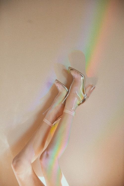 Woman's legs and feet wearing clear, high-heeled boots, legs up against a wall with a rainbow light effect. Fashion and style concept.
