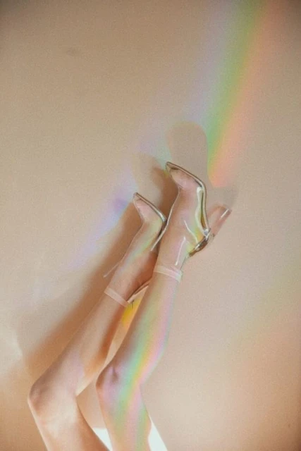 Woman's legs and feet wearing clear, high-heeled boots, legs up against a wall with a rainbow light effect. Fashion and style concept.