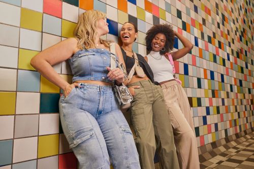 Three women of mixed ethnicities laugh while standing against a patterned tile wall. They are wearing casual tops and pants.