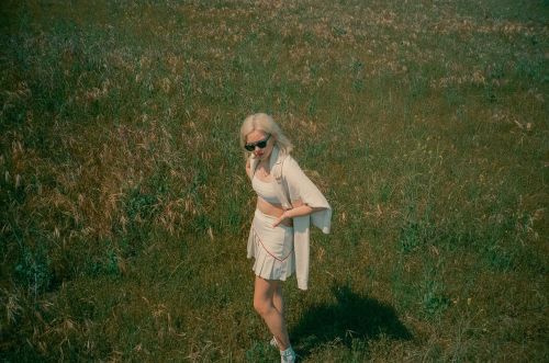 Blonde woman in sunglasses standing in a grassy meadow wearing light-colored crop top and pleated skirt