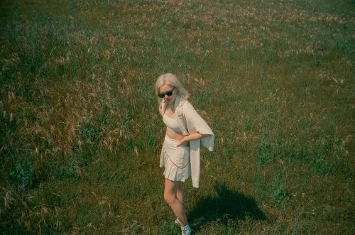 Blonde woman in sunglasses standing in a grassy meadow wearing light-colored crop top and pleated skirt