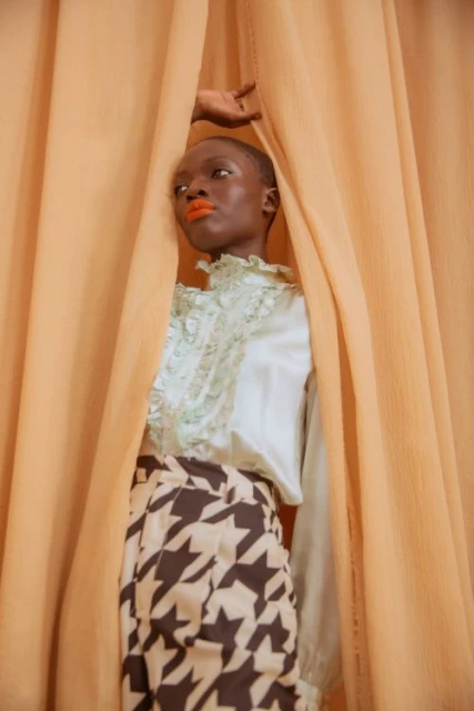 Adult female with buzzcut poses between tan-colored curtains wearing a ruffled blouse and patterned skirt in a portrait composition.