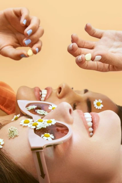 Two women smile with daisy accents on their faces and retro sunglasses, while disembodied hands above them hold small yellow pills.