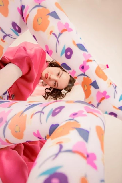 Woman in pink top poses, her legs covered in patterned tights, against light cream background, for a fashion forward editorial shot.
