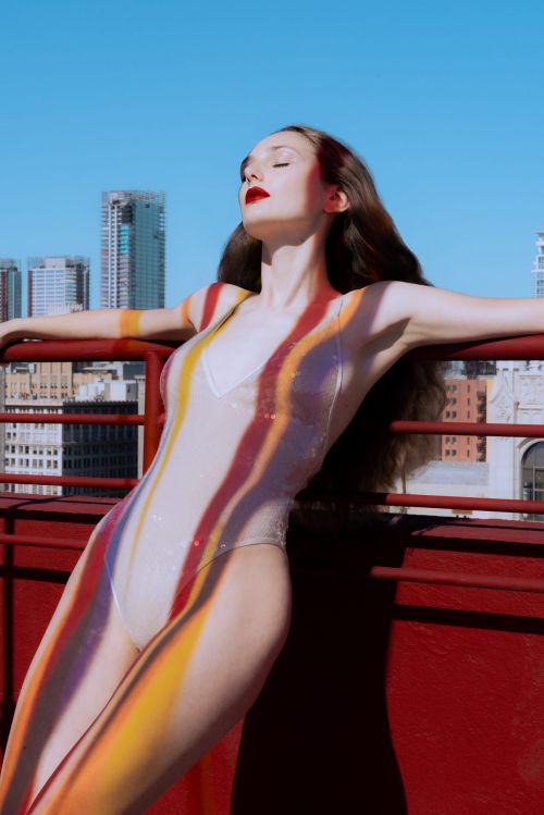 Woman in body paint poses on a rooftop with city skyline in the background; sunny day, clear sky, leaning on red railing.