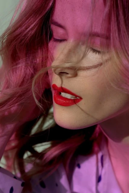 Close-up portrait of a blonde woman with pink hair and red lipstick, eyes closed, wearing a purple shirt, and bathed in pink light.