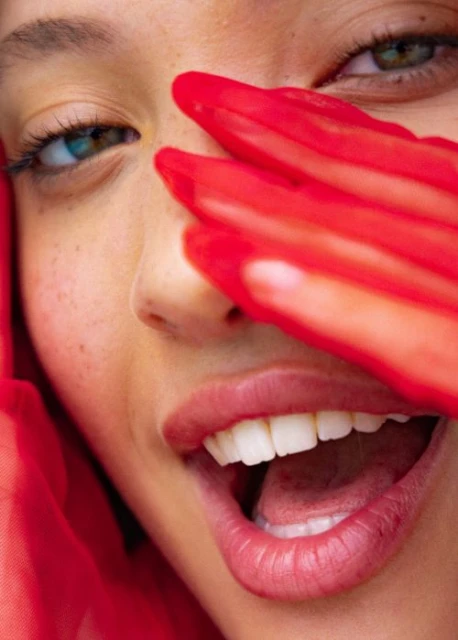Close-up of a young adult woman with heterochromia wearing a sheer red glove over half of her face and smiling with an open mouth.
