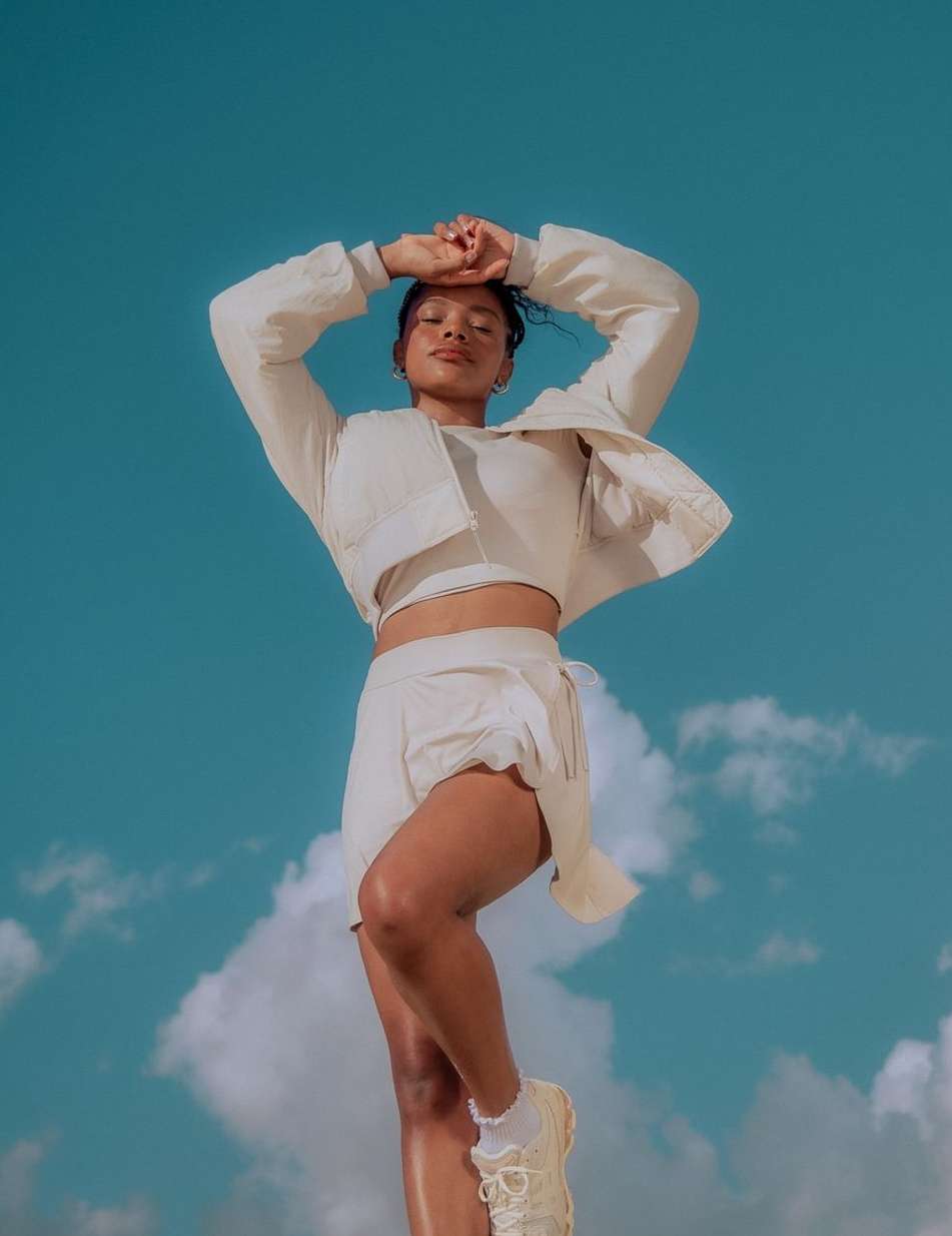 Woman in a cream-colored athleisure outfit strikes a pose with arms raised against a vibrant blue sky with soft, white clouds.