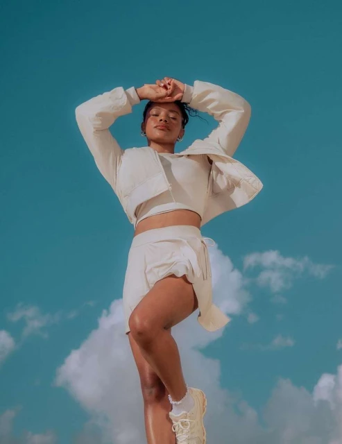 Woman in a cream-colored athleisure outfit strikes a pose with arms raised against a vibrant blue sky with soft, white clouds.