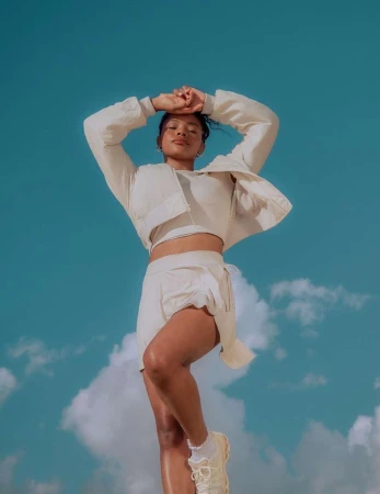 Woman in a cream-colored athleisure outfit strikes a pose with arms raised against a vibrant blue sky with soft, white clouds.
