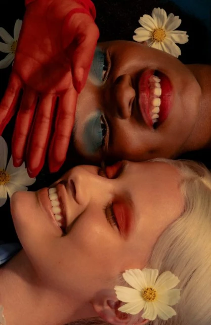 Two women with bold makeup and daisies, one woman is fair-skinned with platinum blonde hair, the other is dark-skinned with black hair.