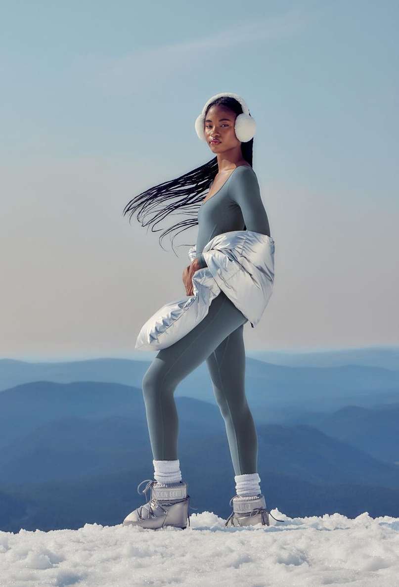 Woman in a grey bodysuit and silver moon boots stands in the snow-covered mountains while wearing white earmuffs in a studio shot.