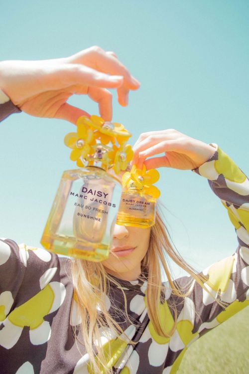 Blonde woman in floral top poses with two Marc Jacobs Daisy perfume bottles, against a blue sky in a bright, sunny setting.