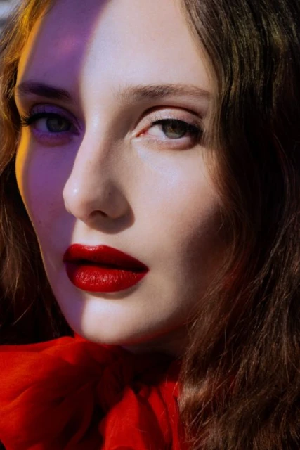 Close-up portrait of a woman with red lipstick, fair skin, and wavy brown hair, featuring dramatic lighting and a red fabric detail.